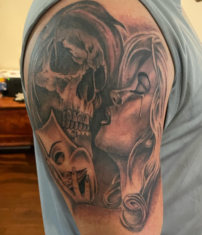 Shoulder tattoo of a skull kissing a woman's face with a mask
